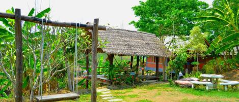 A rustic outdoor area with a swing set, a thatched-roof structure, and a stone pathway.
