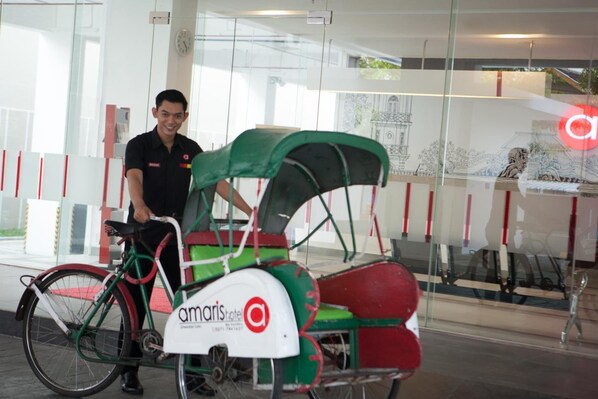 A man standing next to a bicycle with a green and red cart, in front of a glass wall with a reflection of a building and a clock. - Amaris Hotel Sriwedari Solo (Surakarta)