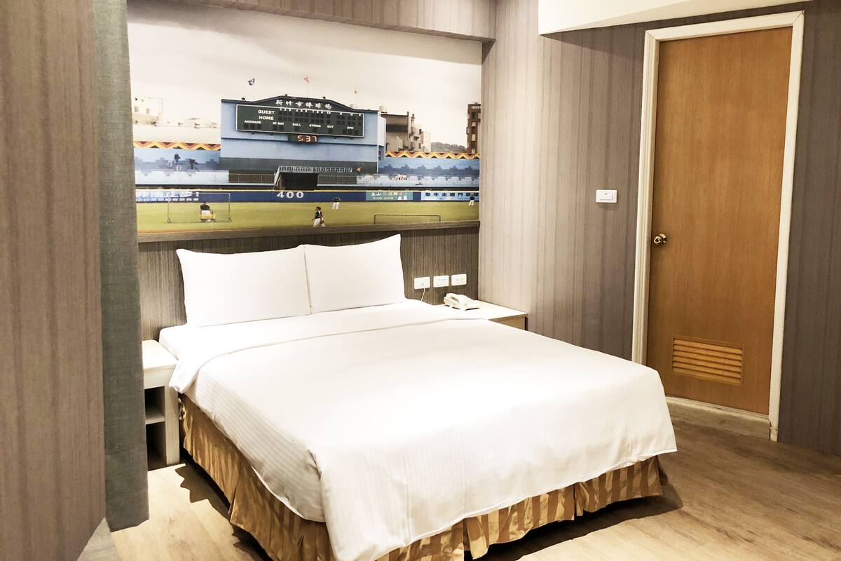 A hotel room with a bed, a nightstand, a wall-mounted TV displaying a sports event, and a wooden door.