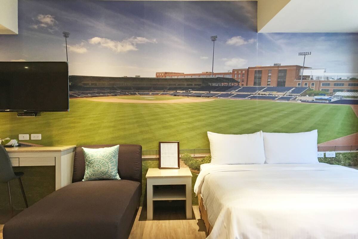 A hotel room with a large mural of a baseball stadium, a bed, a sofa, a nightstand, and a TV.