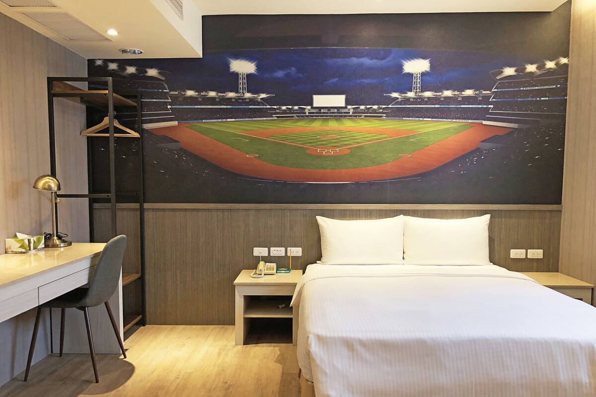 A hotel room with a bed, a desk, a chair, a nightstand, and a wall-mounted TV displaying a baseball field.