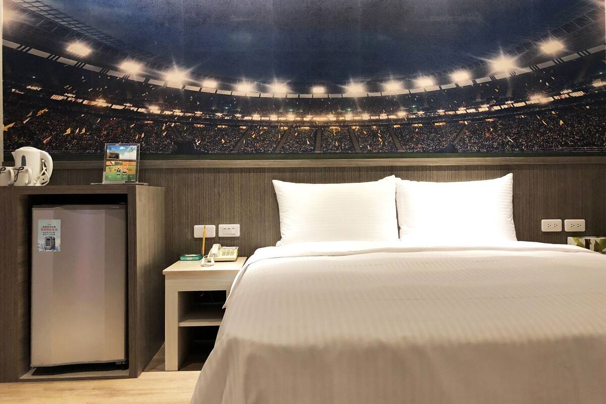 A hotel room with a bed, a nightstand, a phone, a small cabinet, and a poster on the wall.