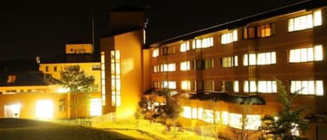 A multi-story building with illuminated windows at night, a flat roof, and a paved courtyard with small trees.