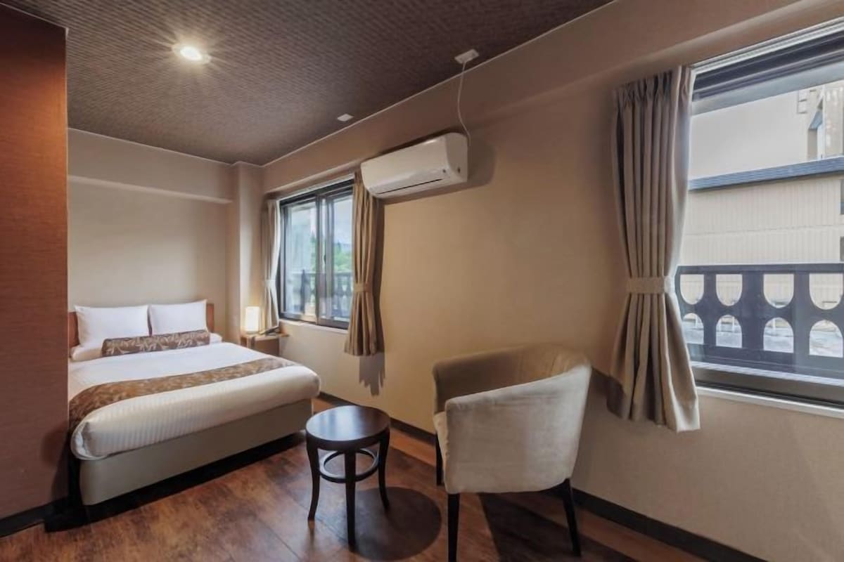 A hotel room with a bed, a chair, a small table, an air conditioning unit, and a window with curtains.