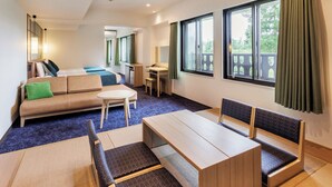 A modern hotel room with a sofa, a dining table, and a small desk. There are large windows with curtains, and a balcony is visible through the windows.