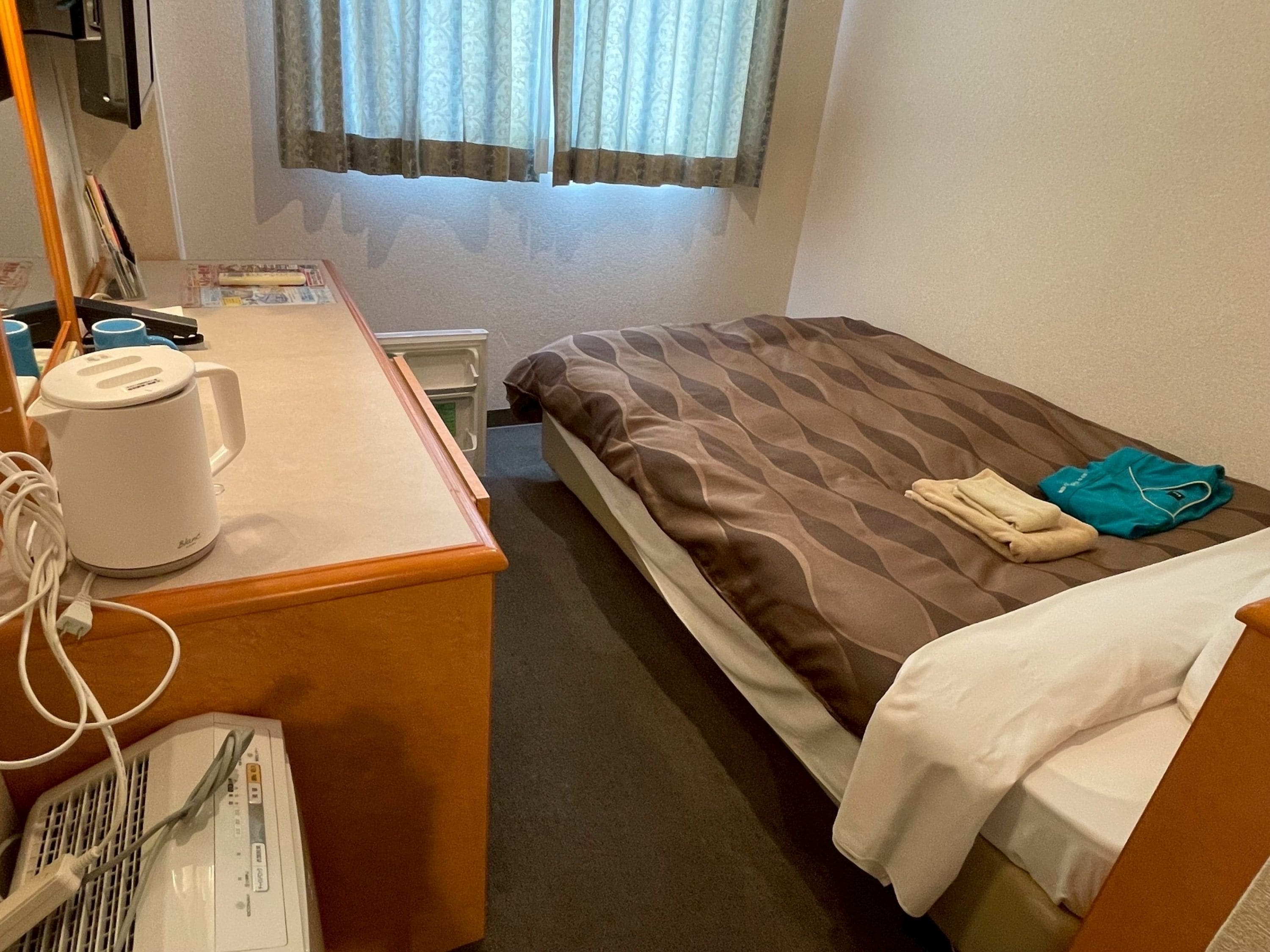 Une chambre d’hôtel avec un lit, un bureau doté d’une bouilloire, une chaise et une fenêtre avec des rideaux.