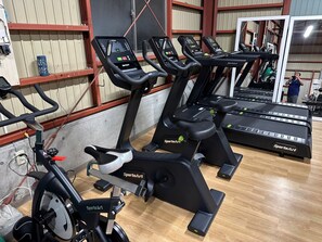 Een sportschool met diverse trainingsapparaten, waaronder loopbanden en hometrainers, met een houten vloer en metalen leuningen.