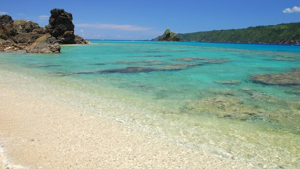 A beach with clear turquoise water, a rocky outcrop, and a distant hill with vegetation. - Hotel Big Marine Amami (Amami)