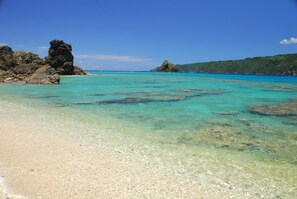 Una playa con aguas turquesas cristalinas, un promontorio rocoso y una colina lejana con vegetación.