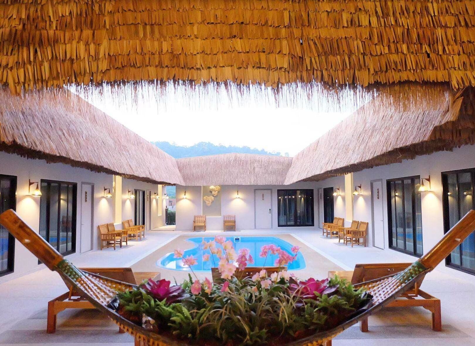 Zona de piscina con techos de paja, tumbonas de madera y vistas a montañas.