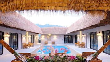 Zona de piscina con techos de paja, tumbonas de madera y vistas a montañas.