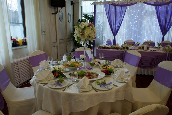 Un comedor con mesas redondas dispuestas para un evento formal, con manteles blancos, cubre sillas morados y centros de mesa florales.
