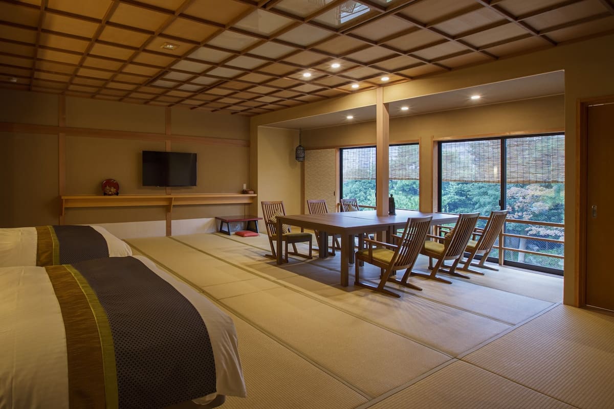 A hotel room with tatami flooring, a dining table with chairs, a TV on the wall, and a view of greenery outside.
