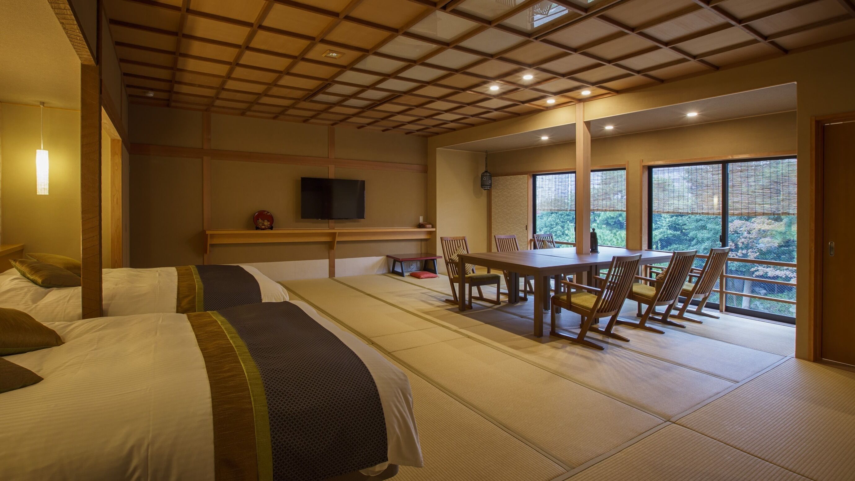Ein Hotelzimmer mit Tatami-Boden, einem Esstisch mit Stühlen, einem Fernseher und Blick ins Grüne durch große Fenster.