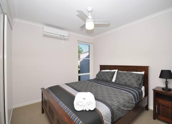 Quarto com cama, ventilador de teto, ar condicionado, janela e mesa de cabeceira com abajur.