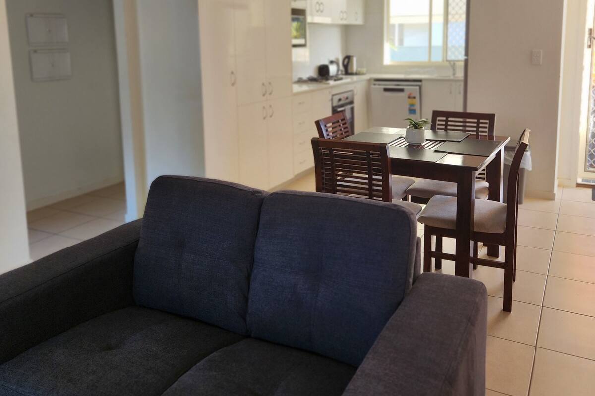 A living room with a dark sofa, a dining area with wooden chairs and a table, and a kitchen in the background.