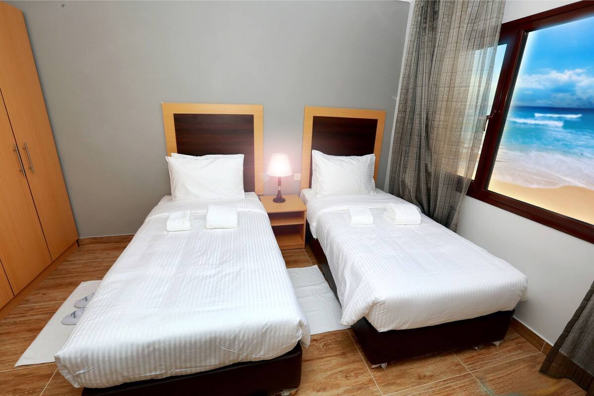 Ein Hotelzimmer mit zwei Einzelbetten, einem Nachttisch mit Lampe, Blick auf den Strand und einem Holzschrank.