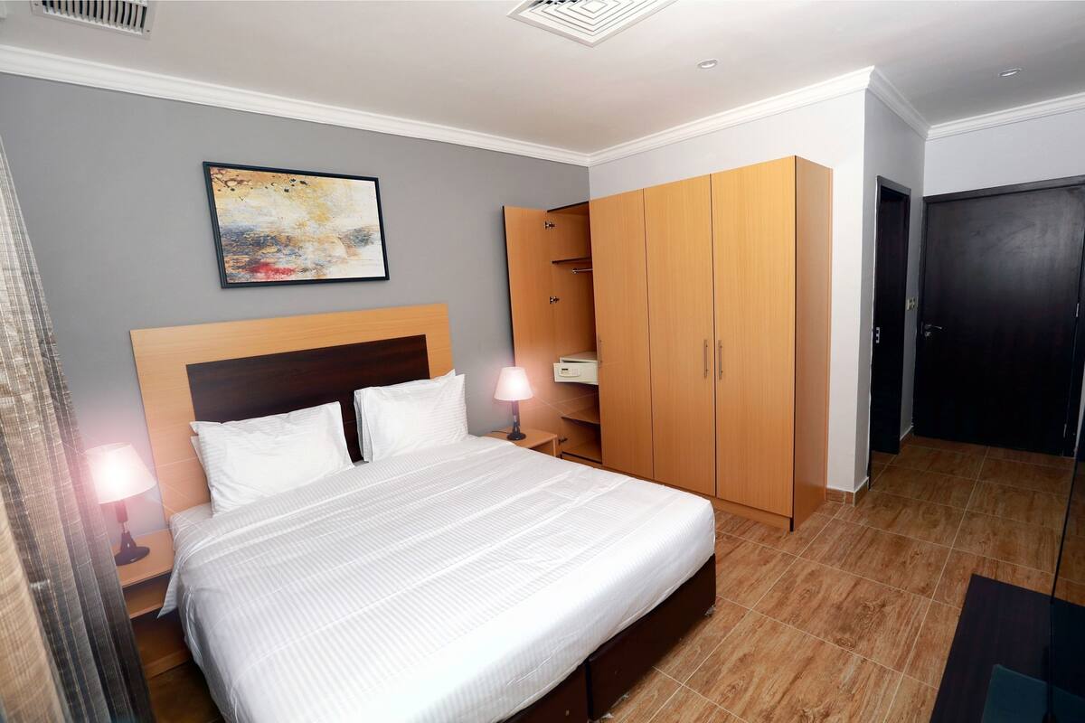 Ein ordentlich eingerichtetes Hotelzimmer mit einem großen Bett, einem Kleiderschrank, einem Nachttisch mit Lampe und einem Bild an der Wand.
