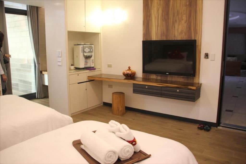 A hotel room with two beds, a TV mounted on a wooden shelf, a coffee machine, and a small trash bin.