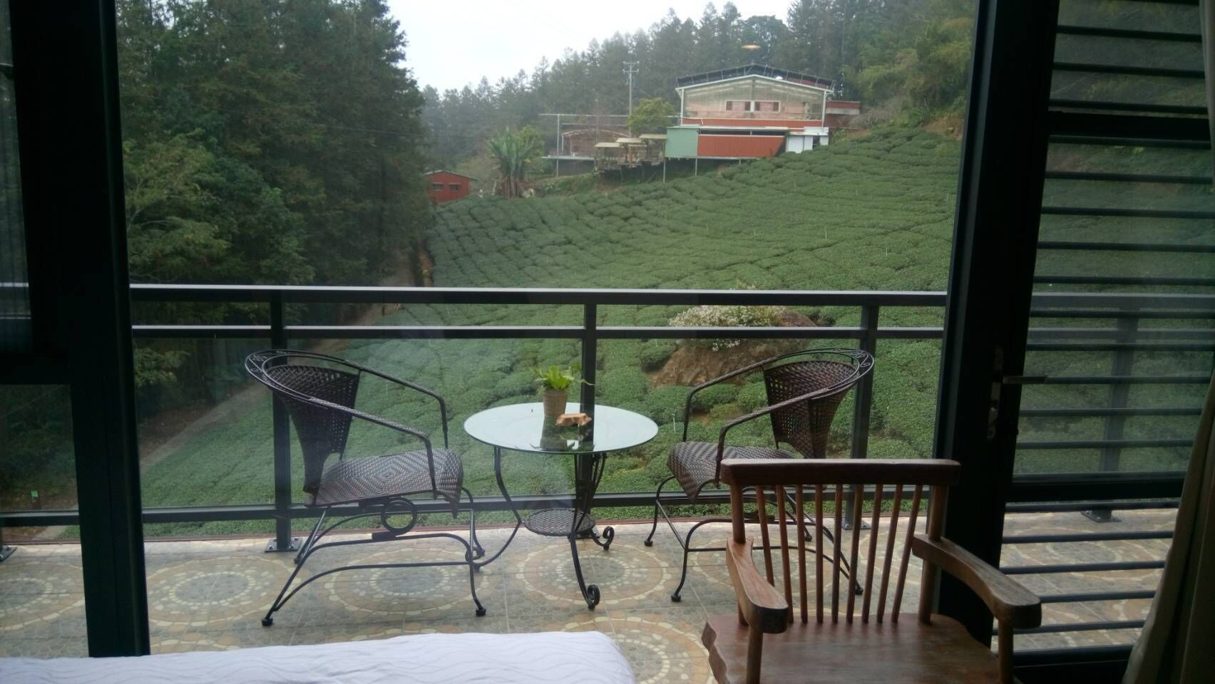 A balcony with a table and chairs, overlooking a tea plantation and a building.