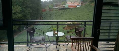 A balcony with a table and chairs, overlooking a tea plantation and a building.