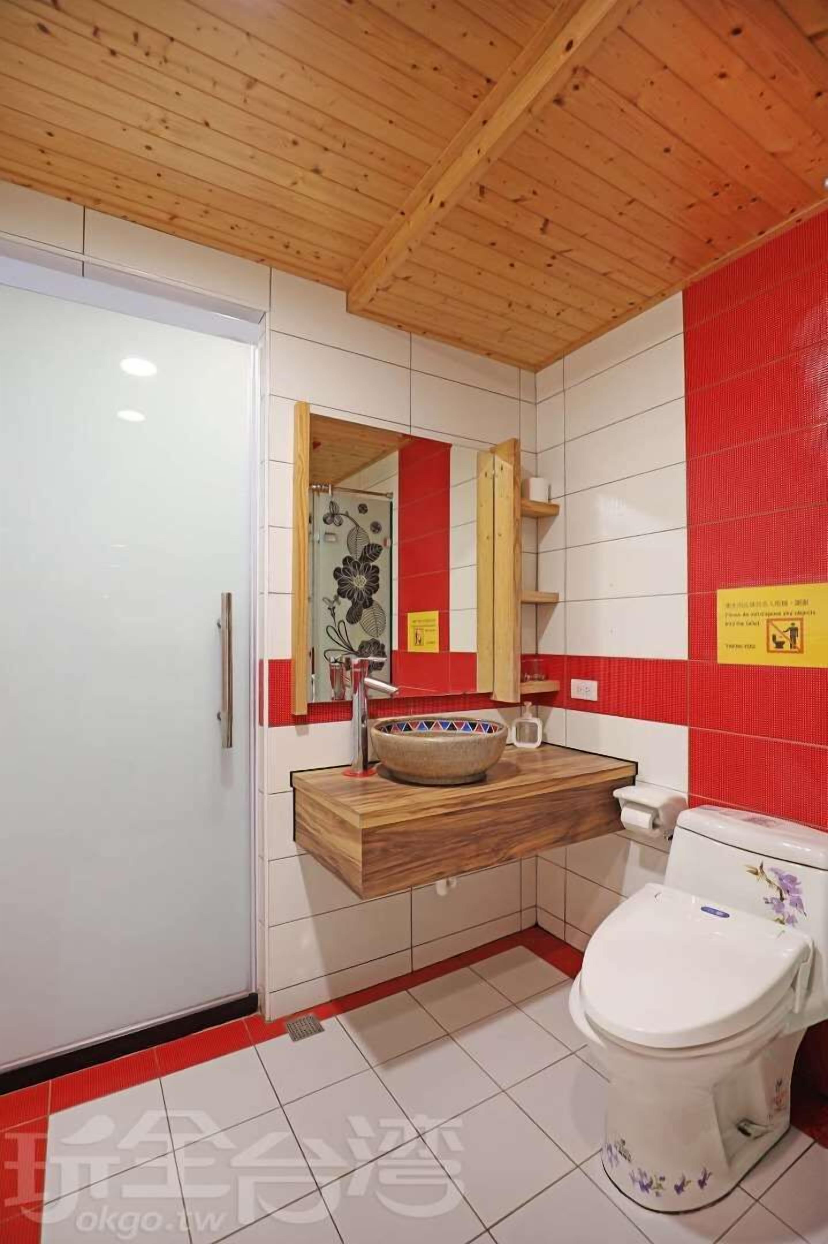 A bathroom with a wooden ceiling, white tiled walls, a red accent wall, a wooden vanity with a bowl sink, a mirror, and a toilet with floral designs.