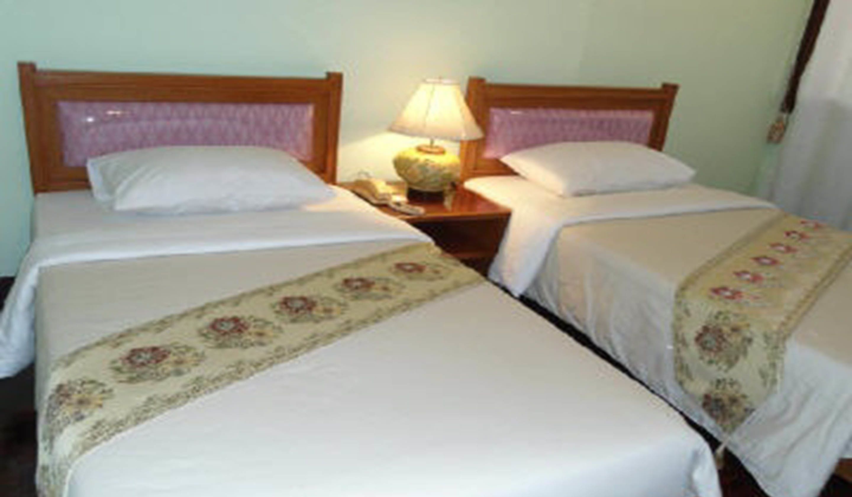 Une chambre avec deux lits simples, chacun doté d’une tête de lit en bois, de draps blancs et d’une couvre-lit à motifs floraux. Une table de chevet avec une lampe se trouve entre les deux lits.