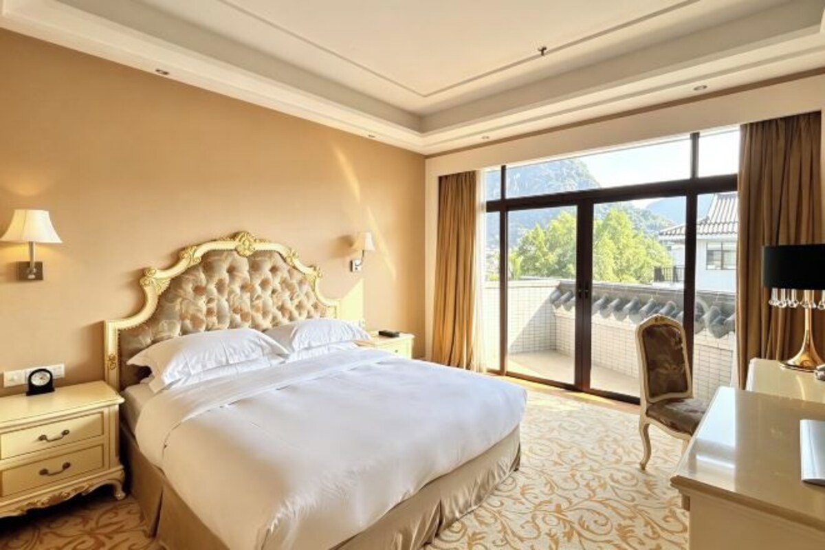 A hotel room with a large bed, a nightstand, a desk, a chair, and a view of the outdoors.