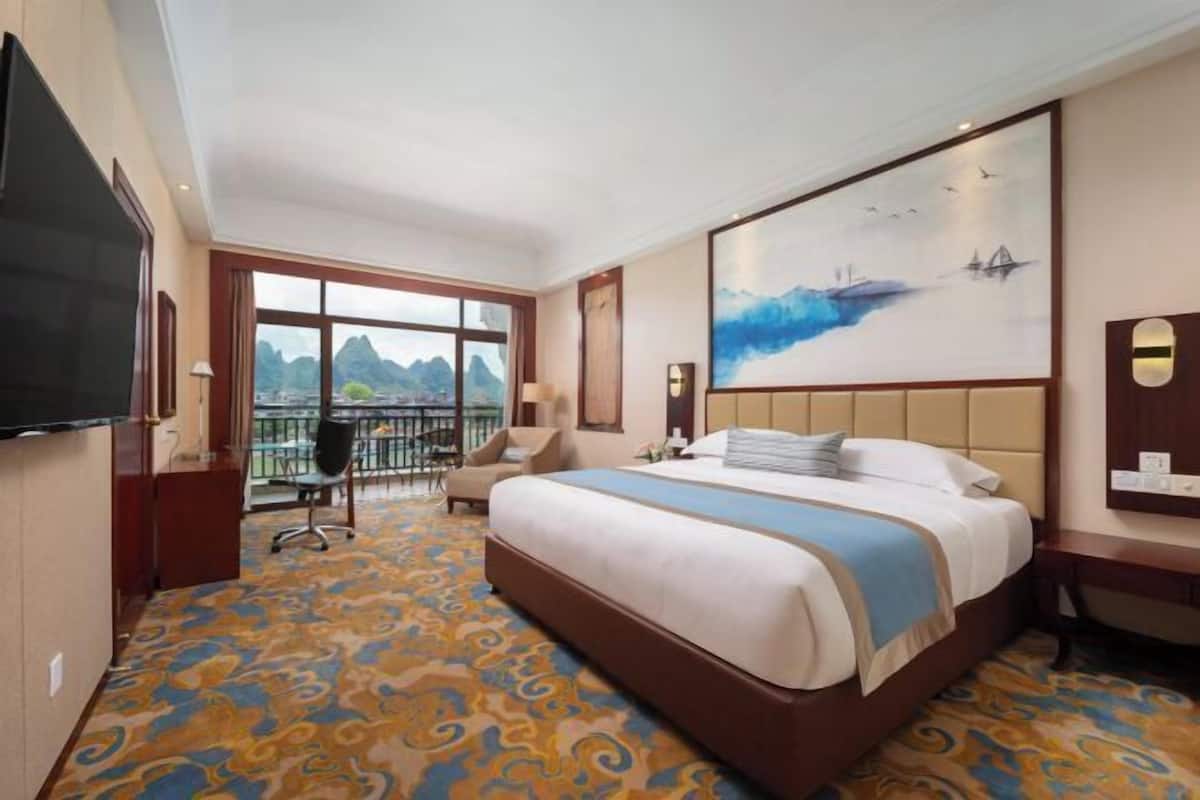 Habitación de hotel con una cama grande, televisión, escritorio y balcón con vistas a las montañas.