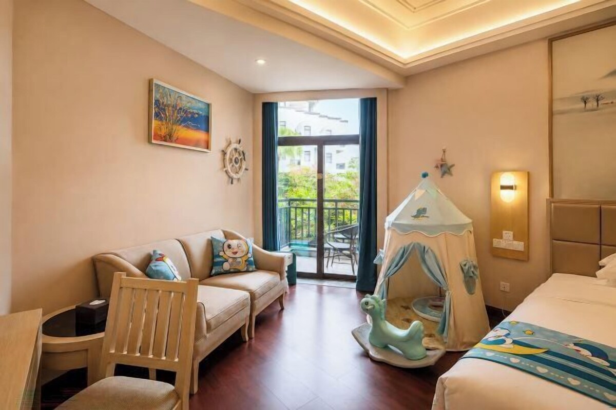 A hotel room with a bed, a sofa, a chair, a small table, a tent-like play area, and a balcony with a view.