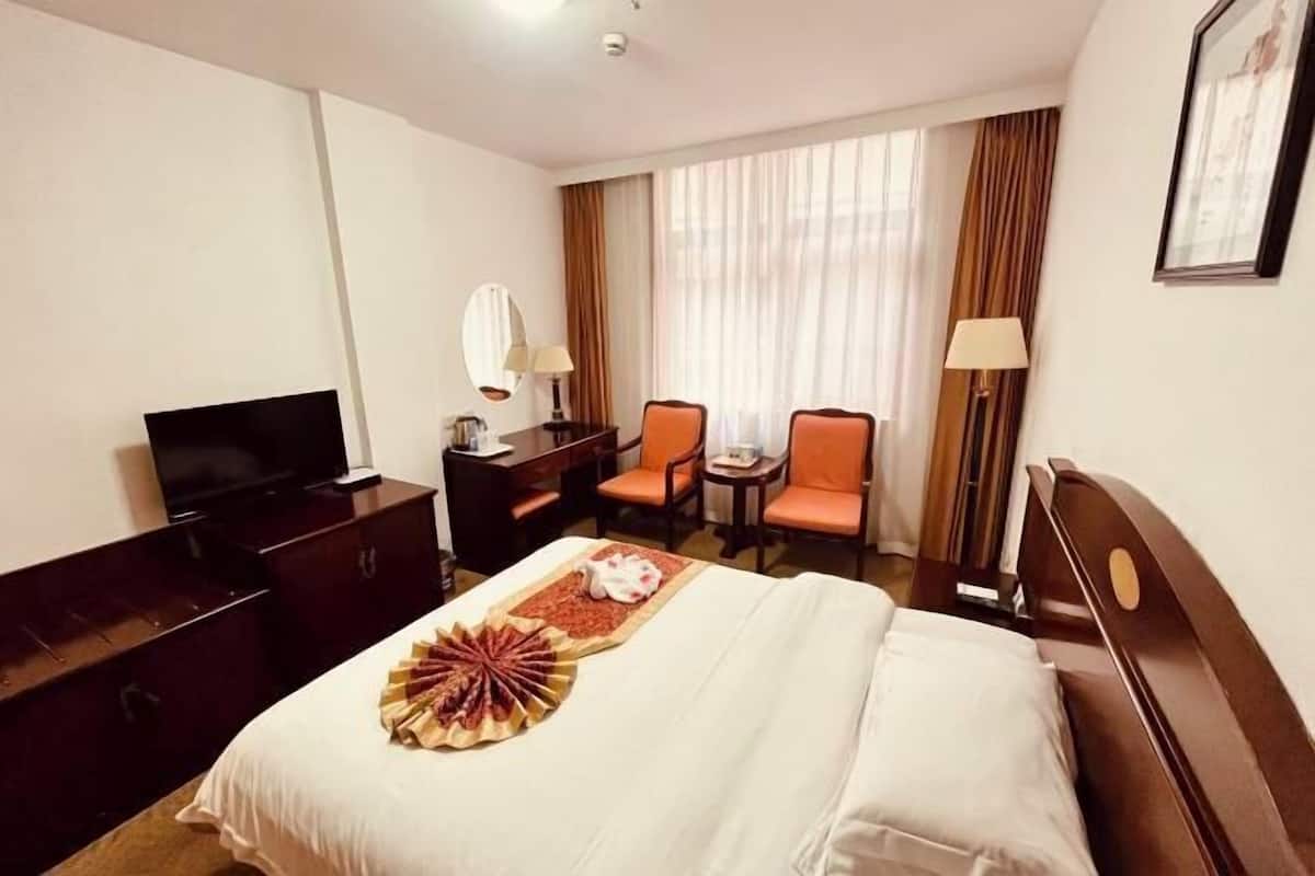 A hotel room with a bed, a TV, a desk, two chairs, and a window with curtains.