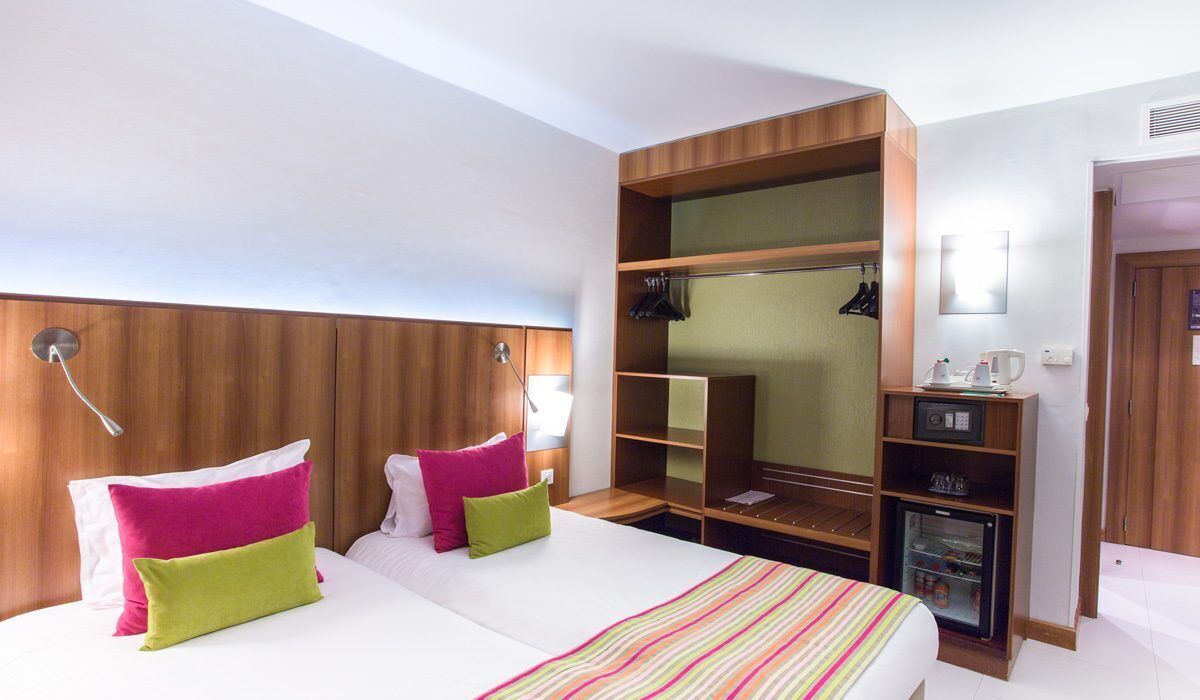 A hotel room with a bed, bedside tables, a wardrobe, a mirror, and a mini-fridge.