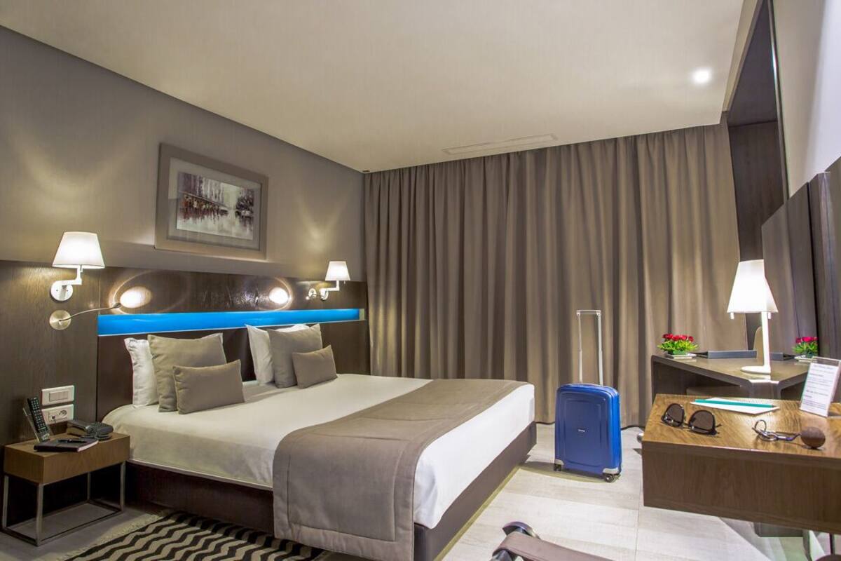 Una habitació d'hotel amb un llit, taules de nit, un escriptori amb un ordinador, un mirall i una finestra amb cortines.