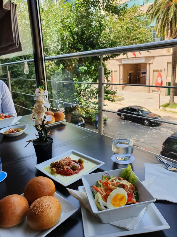 A dining table with food, a potted plant, and a view of a street with cars and buildings. - Le Belvedere (Tunis)