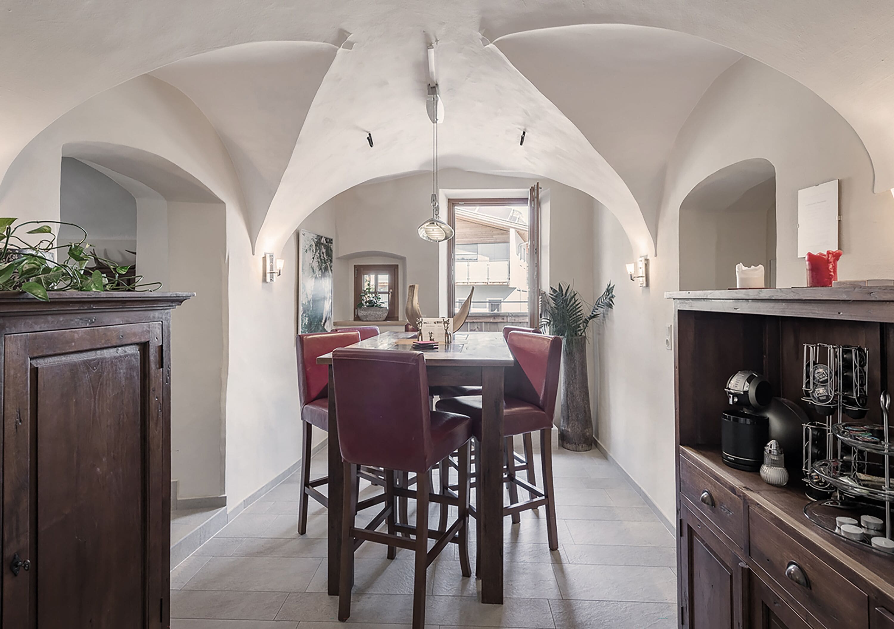 Un comedor con techo alto en arco, una mesa central con sillas y una zona de cocina con electrodomésticos y un armario.