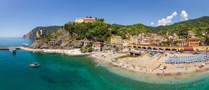 Un pueblo costero con playa, barcos y un edificio parecido a un castillo en una colina.
