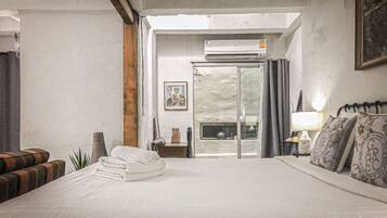 A hotel room with a bed, a small table with a lamp, a window with curtains, and an air conditioning unit.