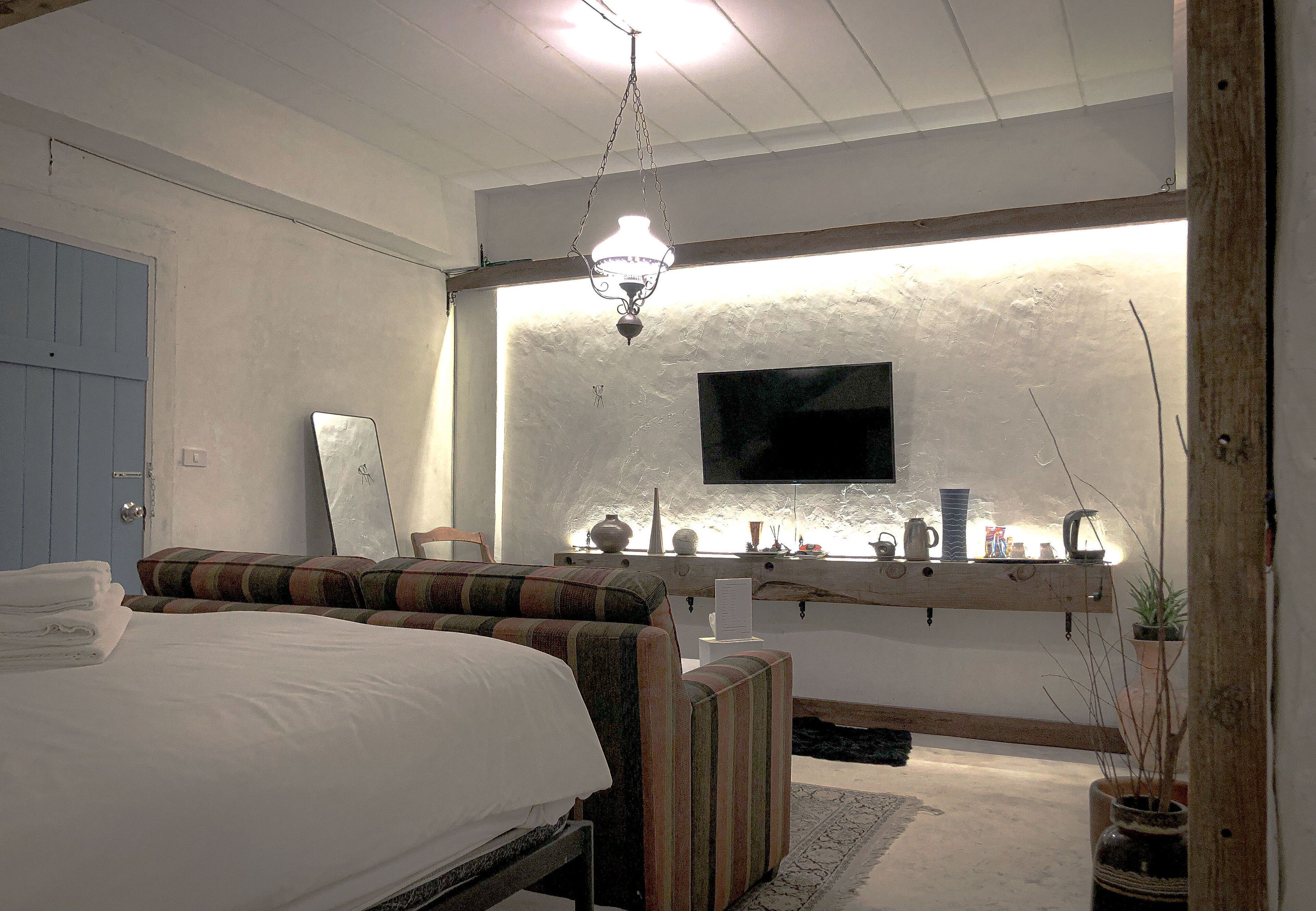A bedroom with a bed, a bedside table, a mirror, a television mounted on the wall, and a shelf with decorative items.