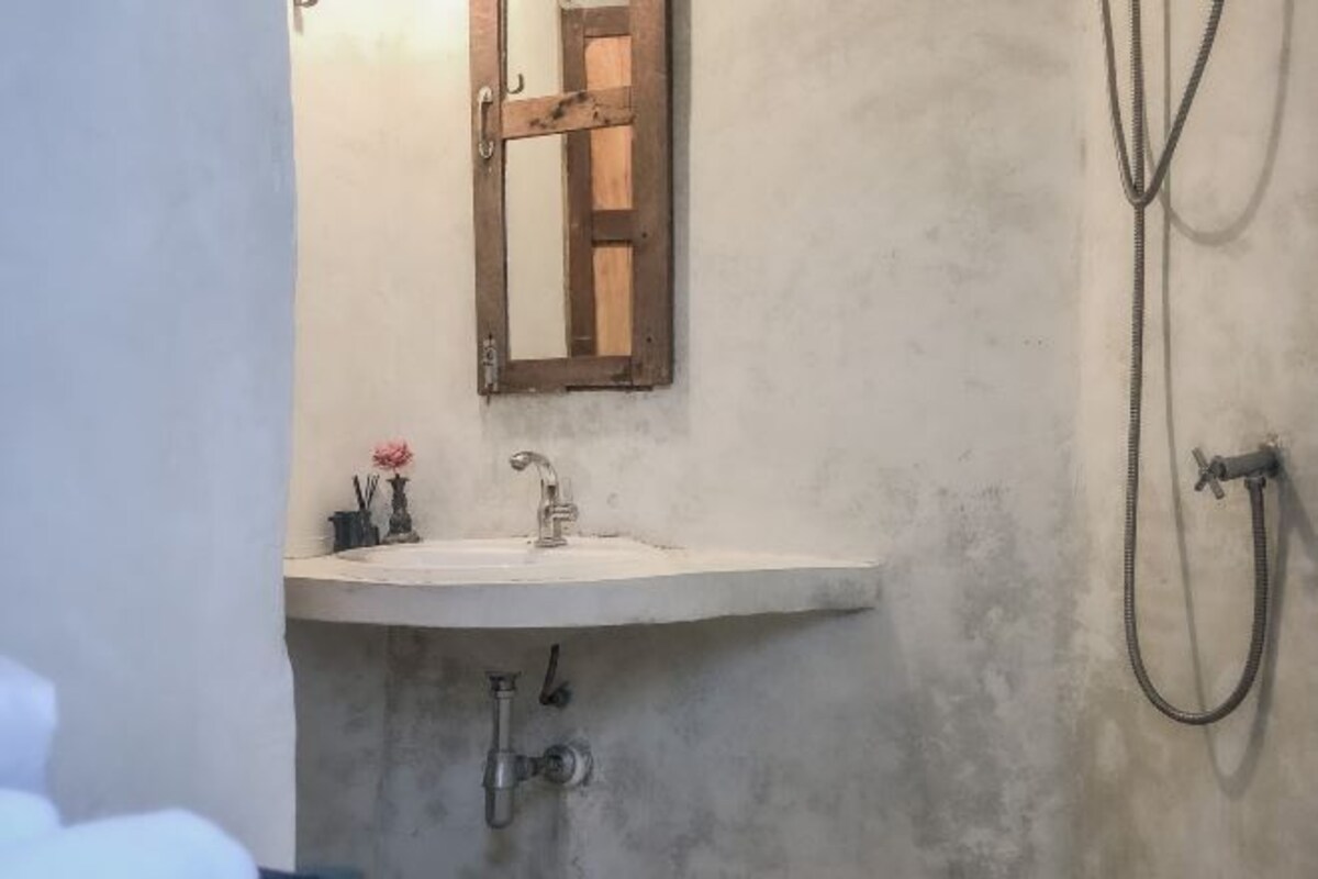 Un baño con un lavamanos blanco, un espejo con marco de madera, un cabezal de ducha de mano y un perchero para toallas.