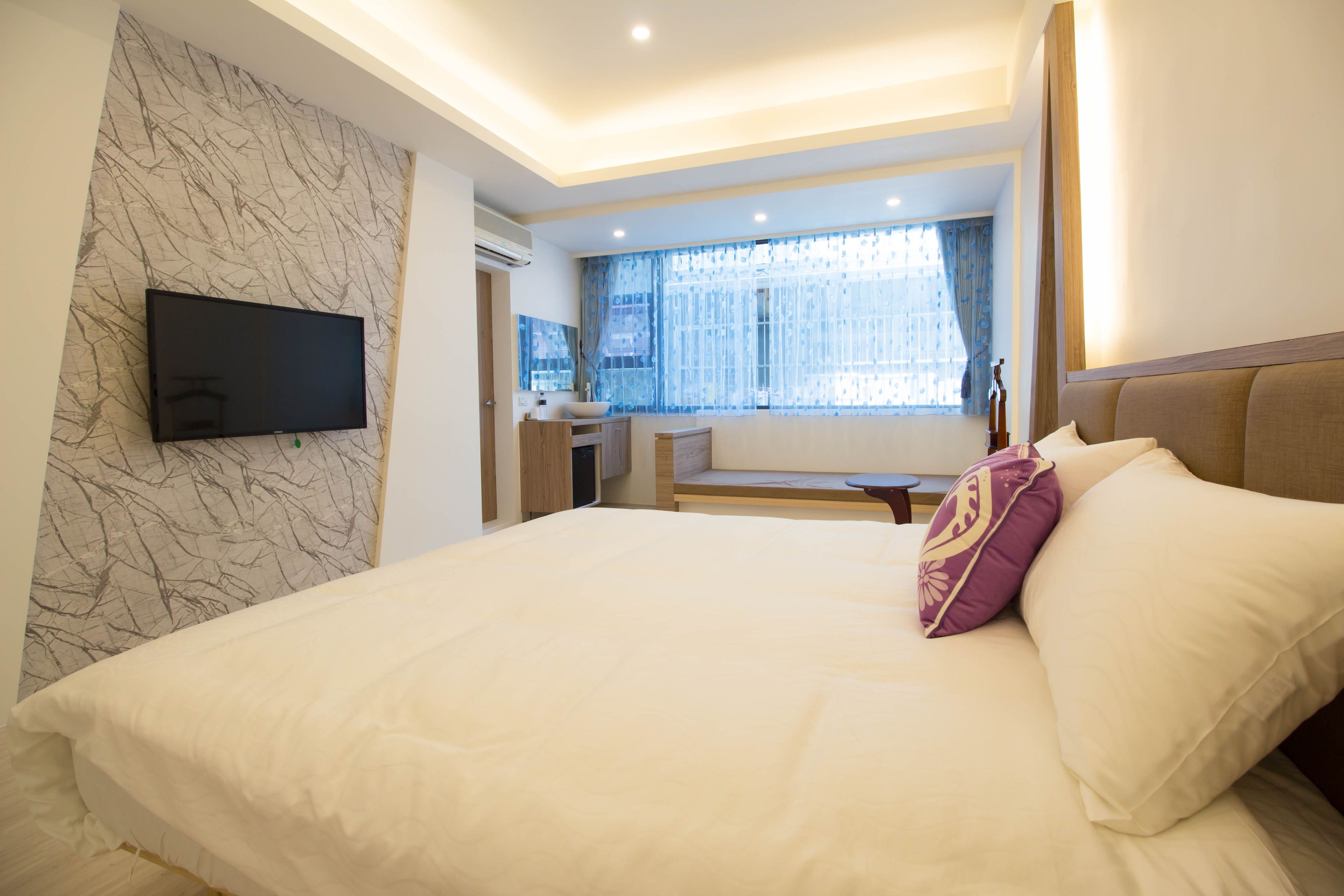 A modern hotel room with a large bed, a flat-screen TV mounted on the wall, and a window with sheer curtains.