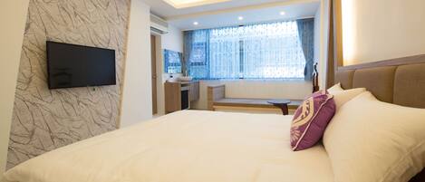 A modern hotel room with a large bed, a flat-screen TV mounted on the wall, and a window with sheer curtains.
