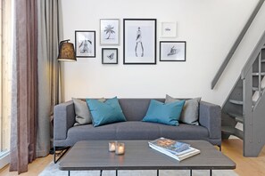 Una sala de estar moderna con un sofá gris, cojines azul verdoso, una mesa de centro con velas y libros, y una escalera a la derecha.