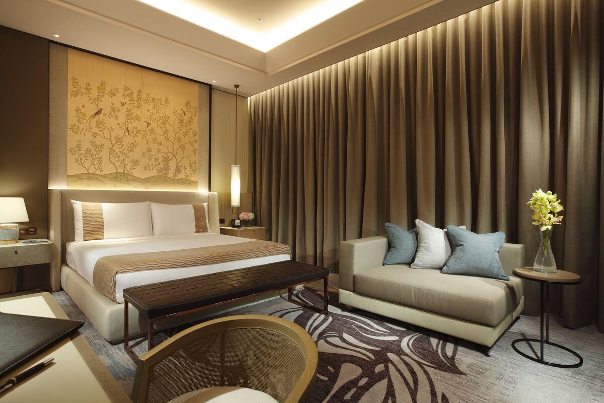A hotel room with a large bed, a sofa, a small table, a chair, and a decorative wall panel.