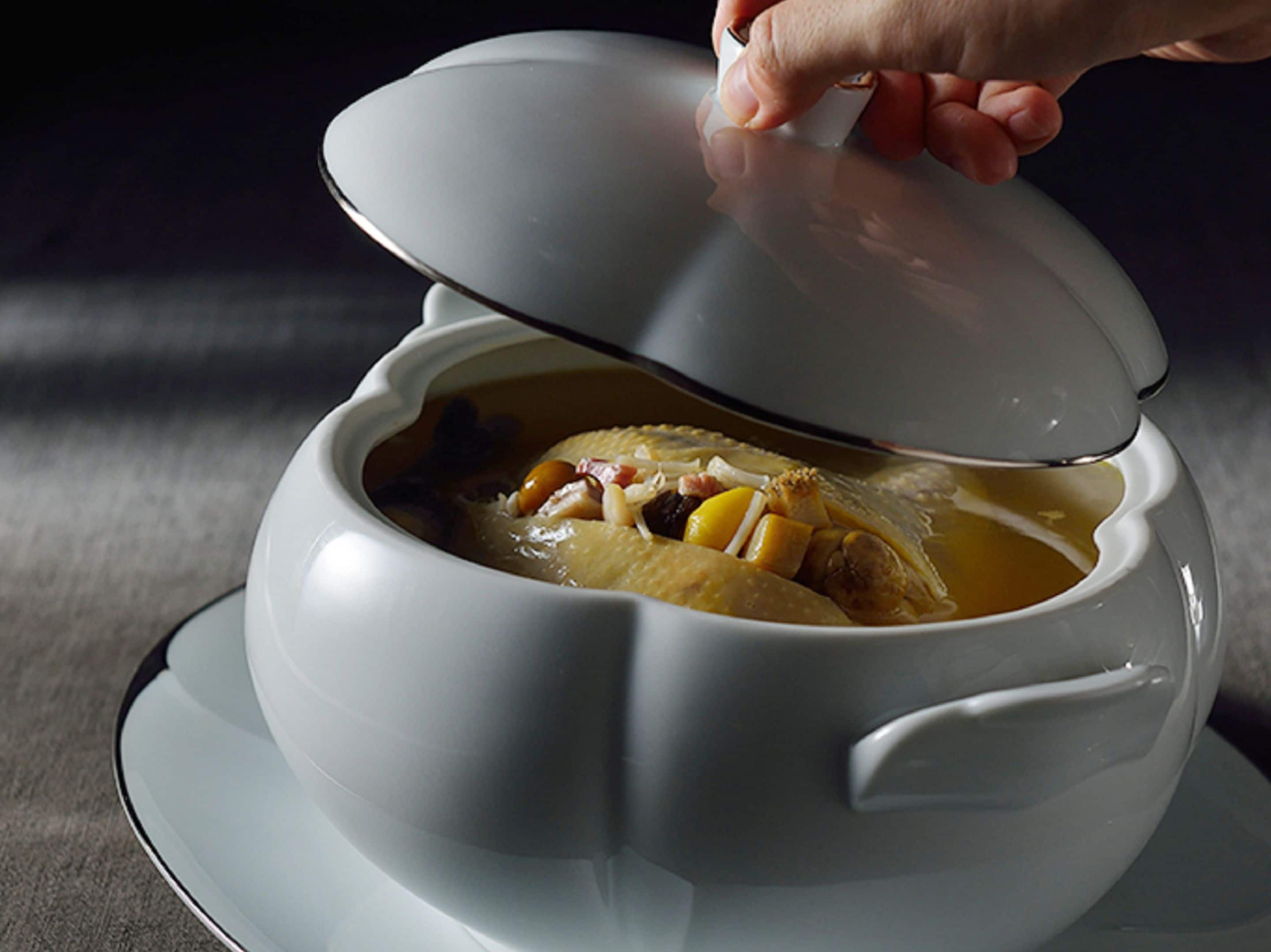 Una olla de cerámica blanca con tapa, mostrando una sopa con ingredientes visibles.