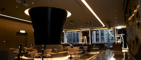A modern lobby with a large black cylindrical sculpture, seating areas, and a view of the cityscape through large windows.