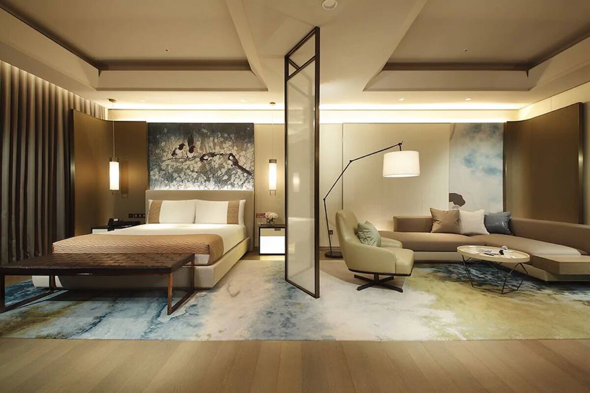 A modern hotel room with a bed, a sofa, a chair, a coffee table, and a large abstract painting on the wall.
