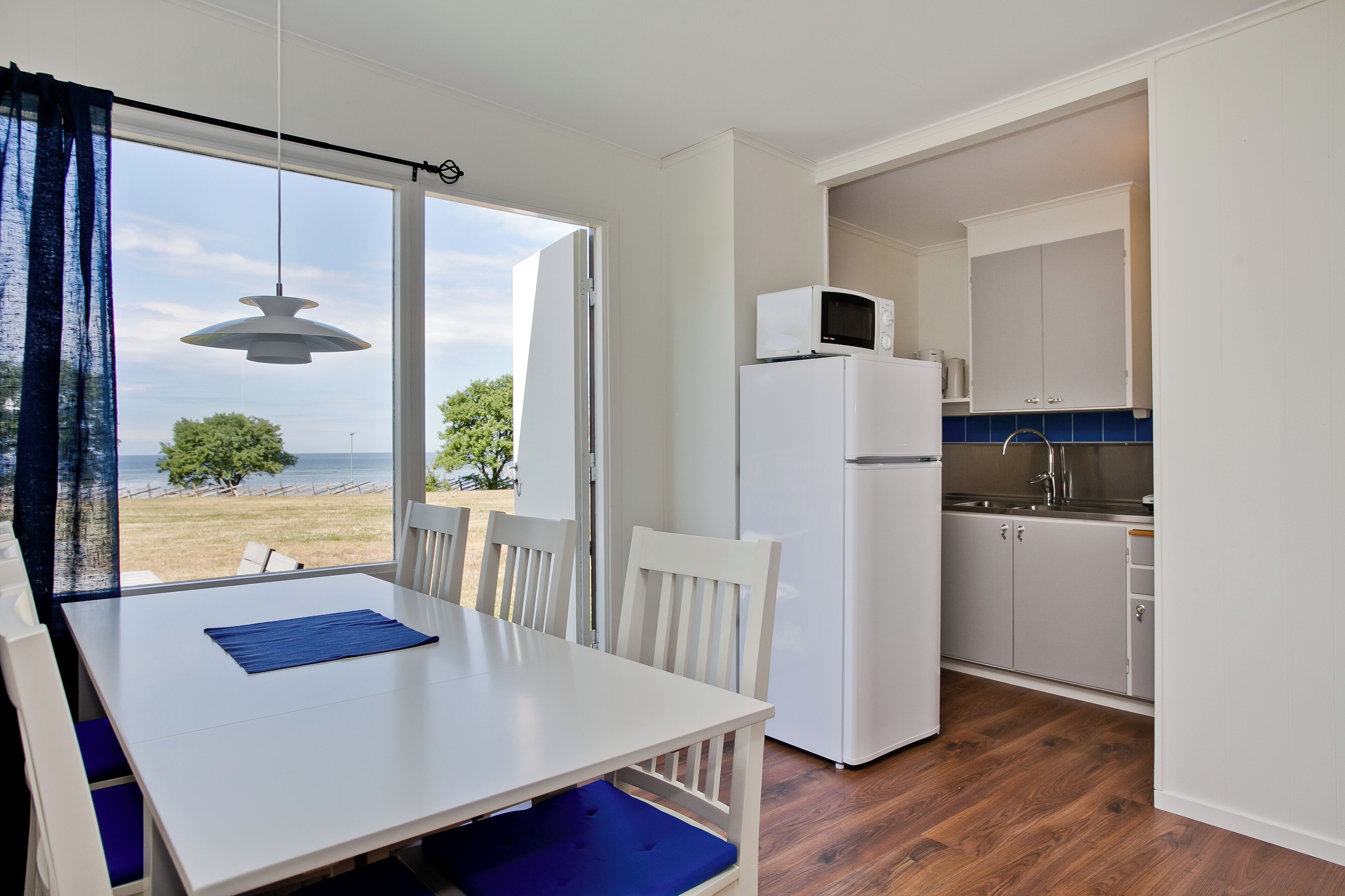 A compact kitchen with white cabinets, a white refrigerator, and a microwave. A dining table with blue cushions is positioned near a large window with a view of the sea.