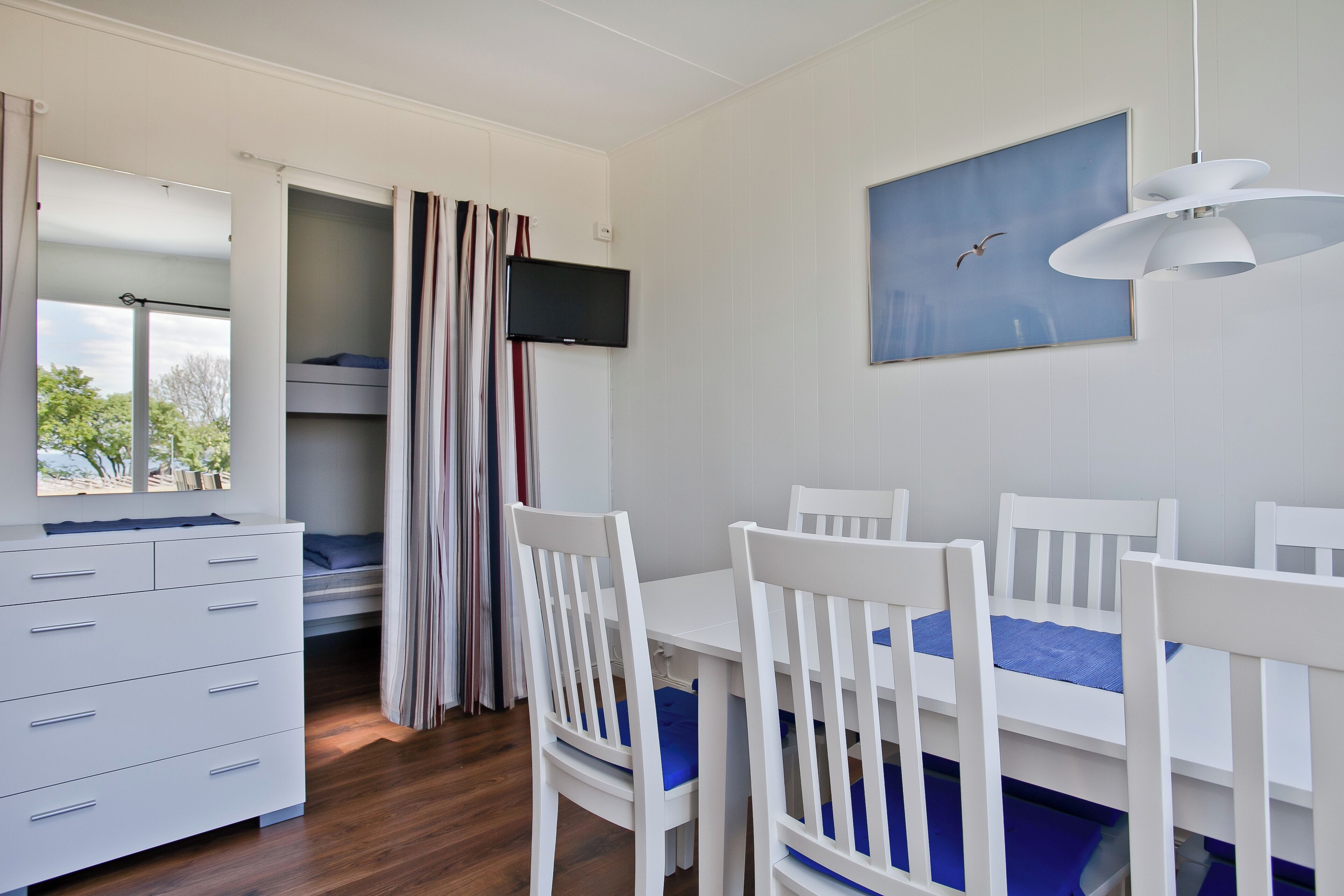 Una habitación con una cama litera, una mesa de comedor con sillas, un aparador blanco, un televisor y un cuadro de pájaros.