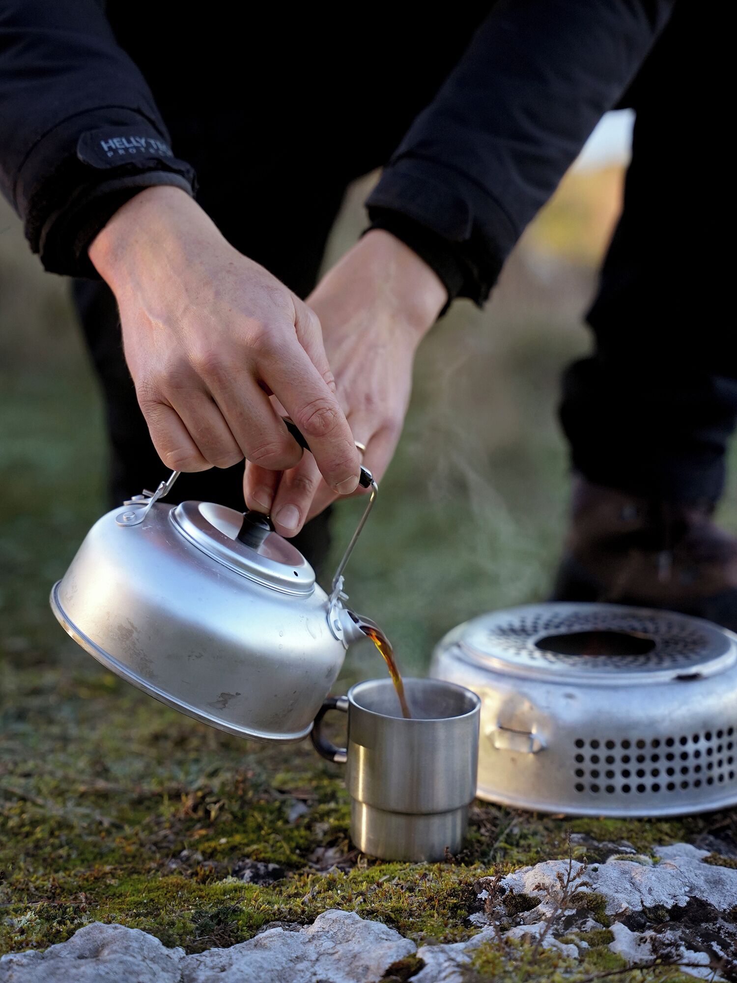 Una persona vertiendo líquido desde un hervidor metálico en una taza metálica sobre una superficie cubierta de musgo.