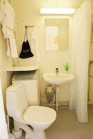 Baño con inodoro, lavamanos y cortina de ducha. Hay un perchero con toallas y una bolsa negra colgada.